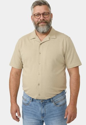 Mittelalter Mann mit grauem Bart und Brille, der ein beiges kurzärmeliges Hemd und blaue Jeans trägt, vor weißem Hintergrund stehend.