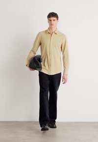 Camisa de manga larga de color beige claro con delante de botones, combinada con pantalones oscuros y zapatos negros. El modelo sostiene un bolso gris. Fondo neutro.