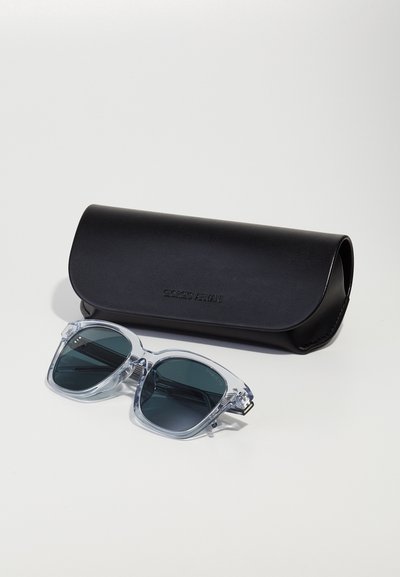 Transparent cat-eye sunglasses with dark lenses, accompanied by a black leather case, featuring embossed branding on the surface.