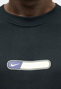 Κοντινό πλάνο ενός ατόμου που φοράει μαύρο πουκάμισο με οριζόντιο οβάλ γραφικό που εμφανίζει ένα μωβ λογότυπο Nike στα αριστερά.