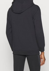 Sudadera con capucha oscura de tejido texturizado, con capucha y bolsillos laterales con cremallera. Presenta un logotipo de marca minimalista en la parte posterior, corte holgado y puños elásticos.