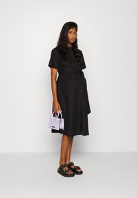 Vestido preto de botões com mangas curtas e cinto na cintura. Modelo segura uma pequena mala lavanda. Sandálias pretas com detalhes de tachas.