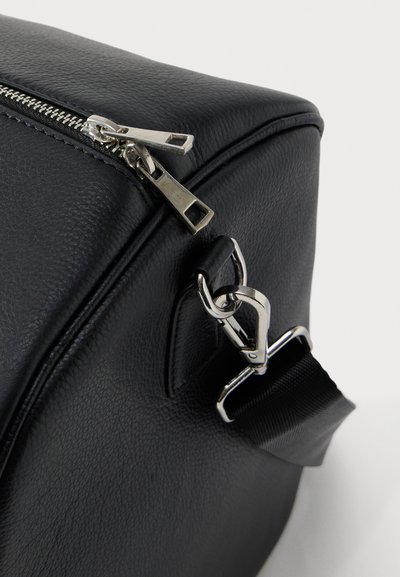 Angolo di borsa in pelle nera con doppia cerniera argentata e gancio per tracolla removibile su sfondo bianco semplice.