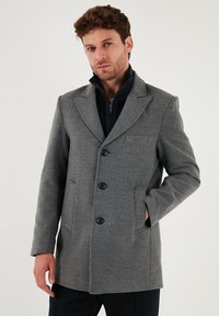 Cappotto di lana grigia con una vestibilità sartoriale, rever a incrocio, chiusura frontale con bottoni e due tasche laterali. Tessuto texture con una finitura liscia.