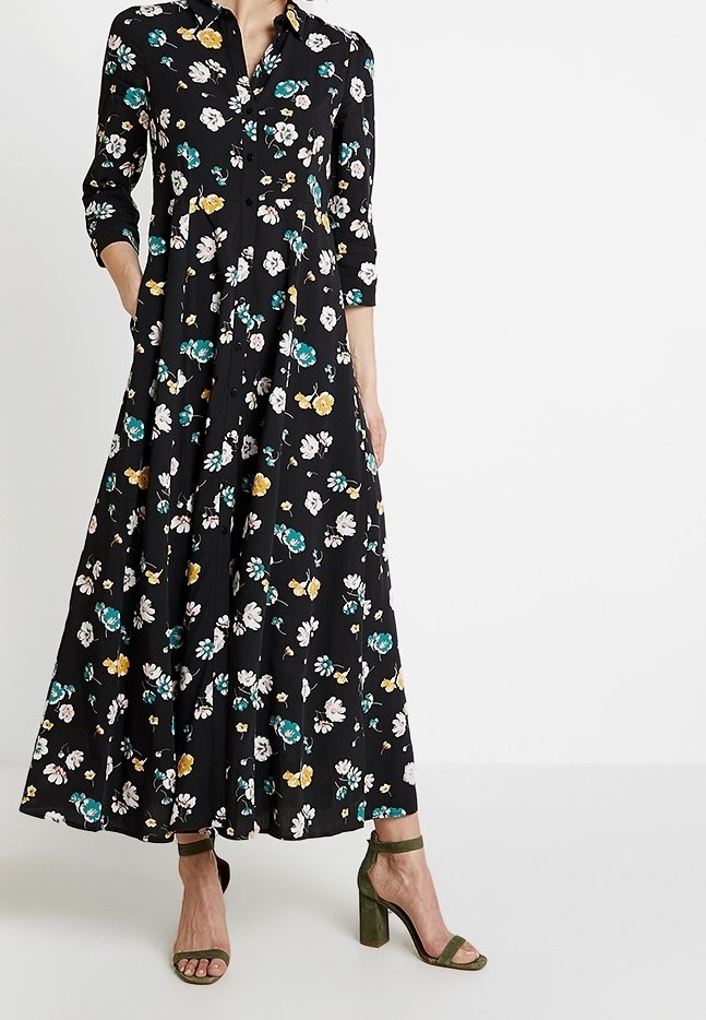 Femme portant une longue robe noire fleurie à manches trois-quarts et des sandales vertes à talons hauts, debout avec une main dans la poche.