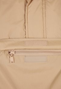 Beżowa pikowana kurtka z kieszenią na zamek, teksturowaną tkaniną i łatą na rzep. Charakteryzuje się subtelnym połyskiem i gładkim wykończeniem powierzchni.