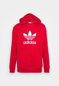 Červená bavlněná mikina s kapucí, s přední kapsou typu klokanka, nastavitelnou kapucí na šňůrku a výrazným bílým logem Adidas se třemi pruhy pod ním.