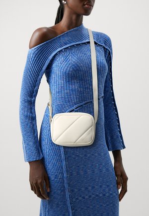Borsa crossbody bianca con motivo a rombi, finitura in pelle liscia e tracolla regolabile, indossata con un vestito blu a coste con bottoni.