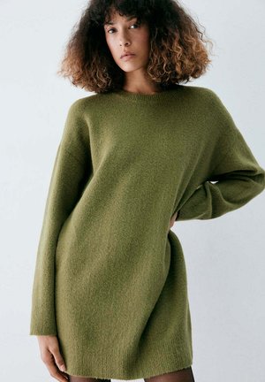 Femme aux cheveux bouclés portant une robe-pull oversize vert olive, debout avec une main sur la hanche devant un fond uni.