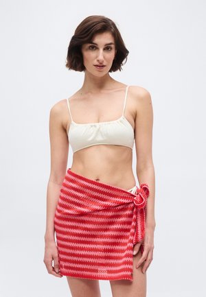 Γυναίκα με κοντά καστανά μαλλιά, που φοράει λευκό τοπ μπικίνι και πλεκτή φούστα με ρίγες κόκκινες και ροζ, δεμένη στη μέση.