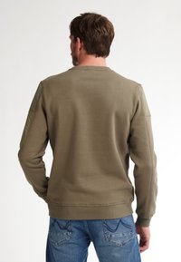 Sudadera verde oliva con textura acanalada, cuello redondo y acentos de hombros acolchados. Lleva puesta con jeans de mezclilla azul.