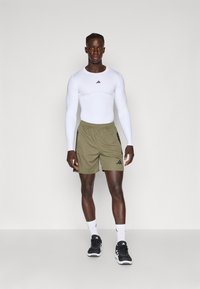 Vit långärmad idrottströja med svart logotyp, olivgröna shorts med svarta detaljer, svarta träningsskor och vita strumpor med logotyp.