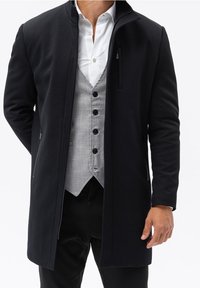 Svart ytterjacka med rak skärning, lättviktsmaterial, dragkedjestängning, sidofickor, lager på en grå väst och vit skjorta.