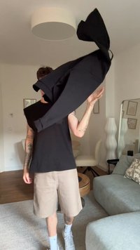 Μαύρο oversized πουκάμισο με μακριά μανίκια και λεία υφή, κρατημένο ψηλά. Οι μπεζ σορτς και τα λευκά αθλητικά παπούτσια ολοκληρώνουν το σύνολο. Εσωτερικός χώρος.