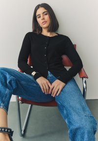 Black long-sleeve ribbed cardigan with buttons, paired with oversized blue denim jeans. Model seated on red chair, hands resting on knees.