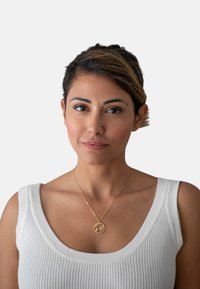 Collana d'oro con un pendente circolare caratterizzato da un design, indossata sopra un top bianco a coste con spalline larghe. Sfondo semplice.