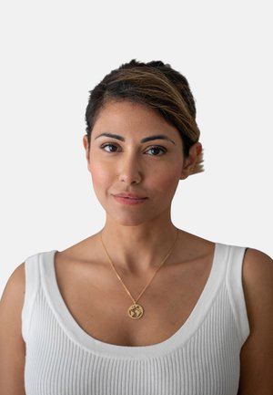 Collana d'oro con un pendente circolare caratterizzato da un design, indossata sopra un top bianco a coste con spalline larghe. Sfondo semplice.