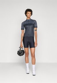 Korte mouwen fietsjersey in donkergrijs met een ritssluiting aan de voorkant, gecombineerd met zwarte shorts. Model houdt een zwarte helm vast; kenmerken zijn gladde texturen.