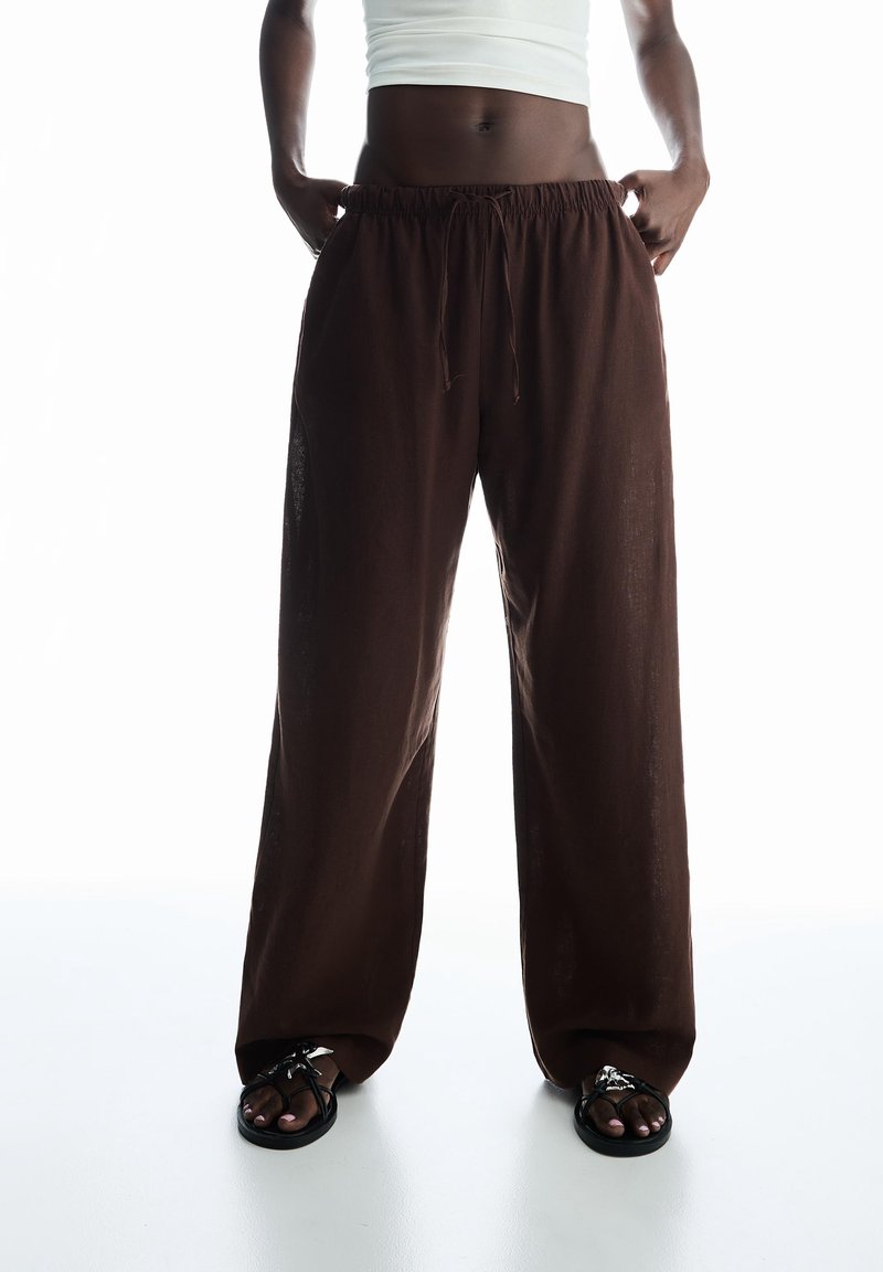 Personne portant un pantalon marron ample avec taille élastiquée à cordon, un haut blanc court et des sandales noires avec un détail en chaîne argentée.