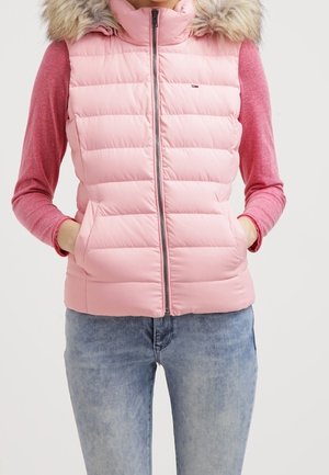 Vrouw draagt een lichtroze doorgestikte bodywarmer met bontkraag capuchon over een lichtroze longsleeve en lichtblauwe spijkerbroek, handen in de zakken van de bodywarmer.