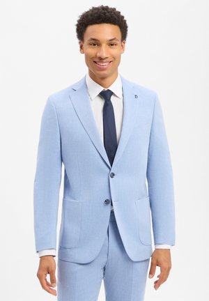 Junger Mann lächelt, trägt einen hellblauen Anzug mit weißem Hemd und dunkelblauer Krawatte, steht vor einem schlichten weißen Hintergrund.