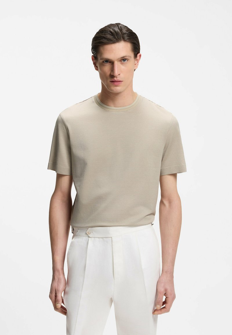 Jeune homme portant une chemise beige texturée à manches courtes et un pantalon blanc taille haute sur un fond blanc uni.