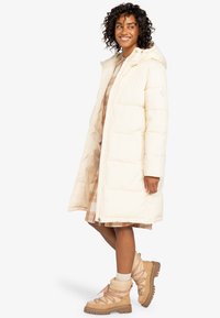 Long manteau en doudoune crème avec capuche et fermeture éclair ; présente un design matelassé. Porté sur une robe à carreaux et associé à des bottes à lacets beiges.