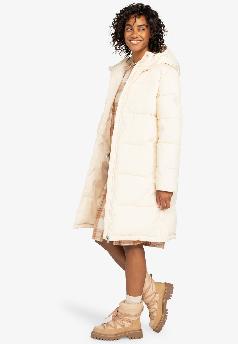 Long manteau en doudoune crème avec capuche et fermeture éclair ; présente un design matelassé. Porté sur une robe à carreaux et associé à des bottes à lacets beiges.