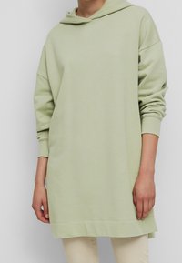 Felpa con cappuccio di colore verde chiaro. Realizzata in un tessuto morbido, presenta spalle scese, maniche lunghe e una tasca a marsupio con un retro leggermente più lungo.