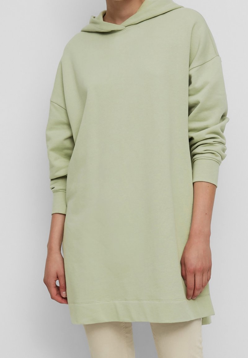 Sweat-shirt à capuche en vert clair. Fabriqué en tissu doux, il présente des épaules basses, de longues manches et une poche kangourou avec un dos légèrement plus long.