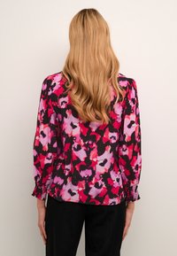 Vrouw met lang golvend blond haar, gekleed in een lange mouwen bloezen met bloemenmotief in roze, rood, zwart en paars, gecombineerd met zwarte broek, staande met haar rug naar ons gekeerd.