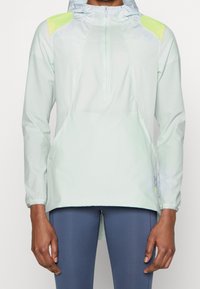 Veste verte clair à manches longues avec une fermeture éclair à l'avant, dotée d'accents jaunes aux épaules et d'une capuche. Fabriquée en matériau lisse et léger.