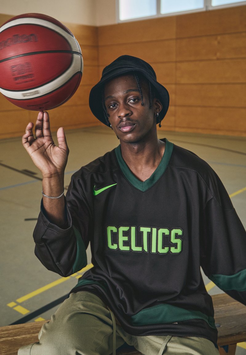 Čierny basketbalový dres s zeleným nápisom "CELTICS", nosený s béžovými nohavicami. Červený basketbal sa otáča na jednej ruke v telocvični.