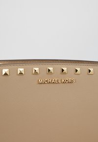Beżowa torebka wykonana z teksturowanego materiału, z złotymi piramidalnymi ćwiekami wzdłuż góry oraz wytłoczonym logo marki "MICHAEL KORS" w złocie.
