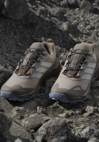 Ζευγάρι μπεζ και γκρι πεζοπορικά παπούτσια Adidas Terrex με ανθεκτικές σόλες σε πετρώδη υπαίθρια περιοχή.