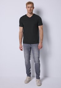 Zwarte v-hals t-shirt, korte mouwen, gecombineerd met lichtgrijze slim-fit jeans. Model draagt beige sneakers, tegen een effen achtergrond.