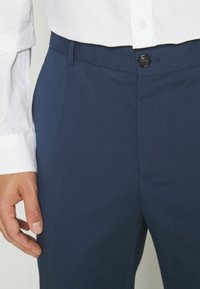 Navy trousers with a smooth texture, featuring a front button closure, belt loops, and a tailored fit. Paired with a white shirt cuff.