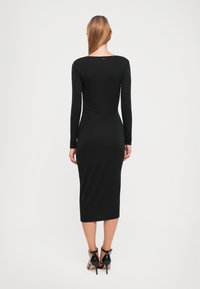 Black long-sleeve midi dress with a fitted silhouette, boat neckline, and smooth texture. Features a subtle back detail and paired with black heels.