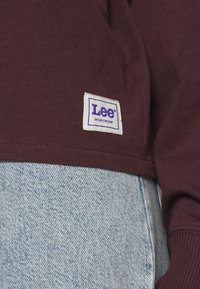 Hnědá košile s dlouhým rukávem s bílou etiketou Lee Workwear. Nasažená přes světle modré džínové kalhoty s hladkou texturou a viditelnými švy.