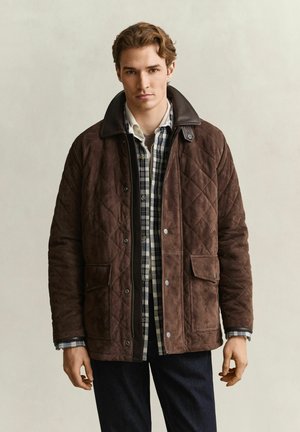 Young man wearing a brown quilted jacket over a black and white plaid shirt, standing against a plain light background.