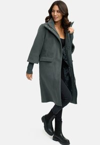 Manteau en polaire vert foncé, long, avec un col haut, une fermeture à boutons sur le devant et des poches latérales, associé à un jean slim noir et des bottines.