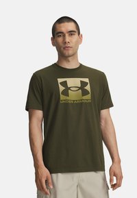 Camiseta de manga corta verde oliva hecha de algodón, con un gran logo de Under Armour en oro y negro en un diseño cuadrado en el frente.