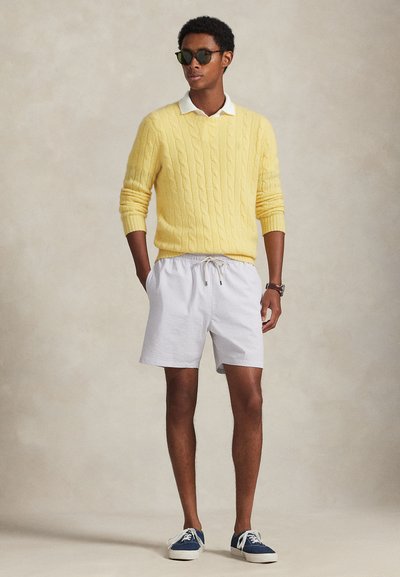 Άνδρας που φοράει κίτρινο πλεκτό πουλόβερ με σχέδιο καλωδίου πάνω από λευκό πουκάμισο με γιακά, ανοιχτόχρωμα σορτς με σχέδιο, γυαλιά ηλίου, ρολόι και μπλε αθλητικά παπούτσια, στέκεται με τα χέρια στις τσέπες.