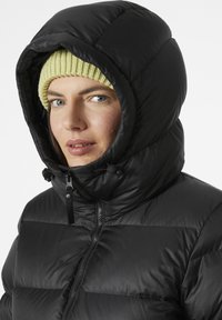 Schwarze Pufferjacke mit hohem Kragen und großer Kapuze. Verfügt über einen Frontreißverschluss und verstellbare Kordeln. Getragen mit einer hellgrünen Strickmütze.
