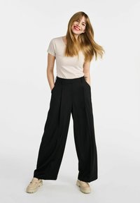 Czarne spodnie typu wide-leg z fałdami z przodu, zestawione z jasnobeżowym, dopasowanym t-shirtem i butami na platformie. Modelka stoi z rękami w kieszeniach.