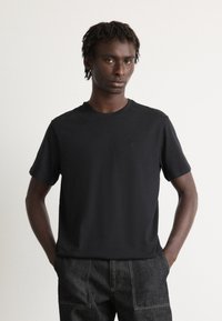 Man met korte dreadlocks die een eenvoudige zwarte t-shirt en donkere spijkerbroek draagt, staande met zijn handen in de zakken tegen een witte achtergrond.
