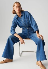 Chemise en denim avec une poche avant, jeans évasés taille haute bleu moyen, et chaussures plates beiges, assis sur une chaise en métal.