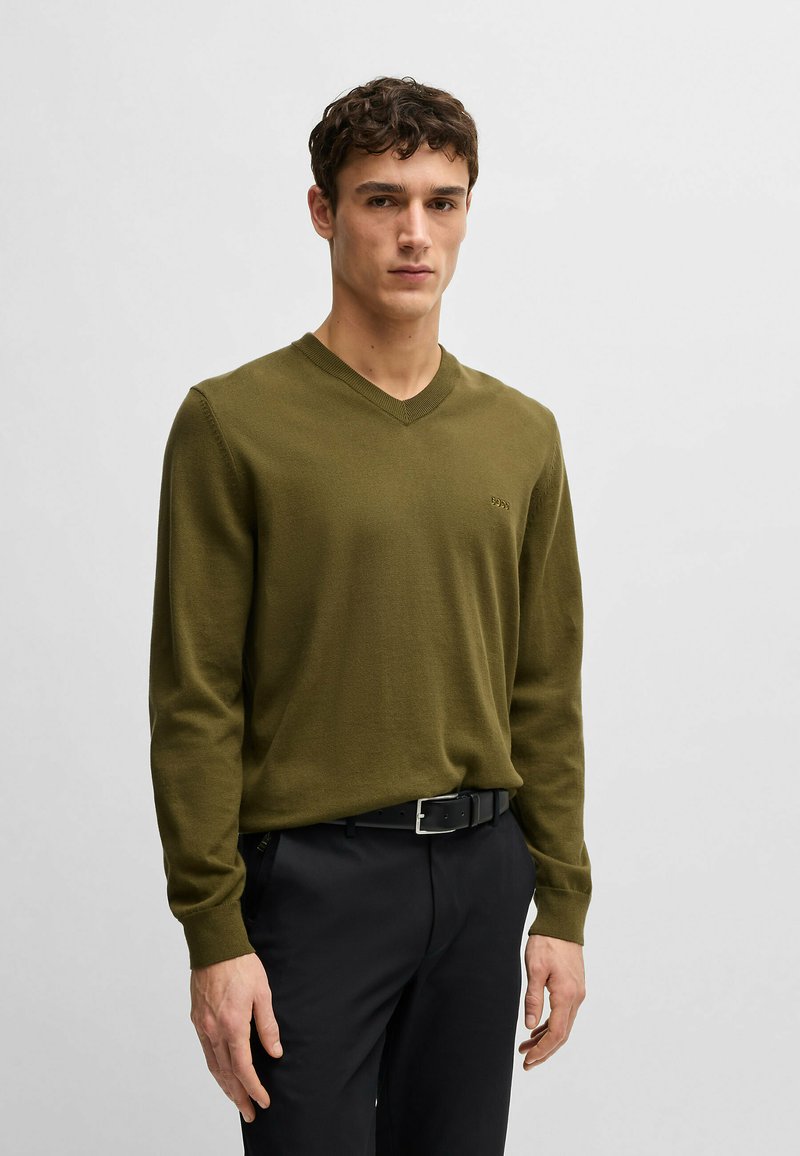 Maglione leggero verde oliva a maglia con scollo a V, maniche lunghe e un logo discreto sul petto, abbinato a pantaloni neri e una cintura.