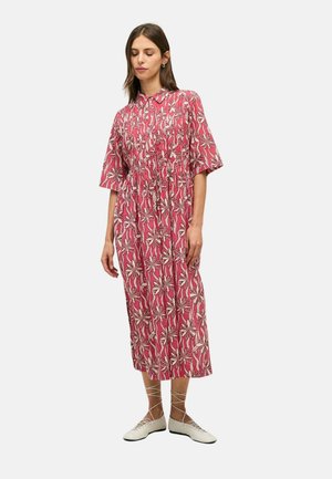Mujer con vestido midi rosa con estampado floral, mangas cortas, cuello con botones y zapatos planos blancos con cordones, de pie frente a un fondo liso.