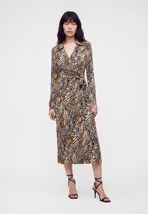 Femme portant une robe portefeuille à manches longues, longueur genoux, avec un motif zèbre abstrait noir, blanc et or, et des talons hauts noirs à brides.